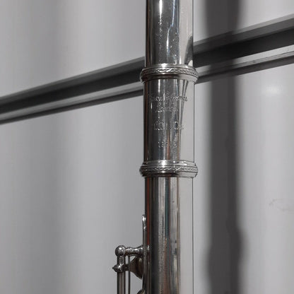 A close-up of a silver flute body with key mechanism, photographed against a light gray background with dark horizontal lines.