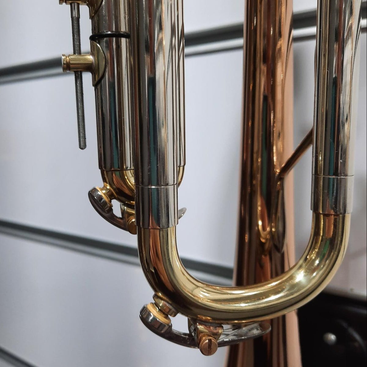 Close-up of a brass instrument, showing shiny gold and copper-toned metal valves, tubing, and bell against a light gray slatted wall background.