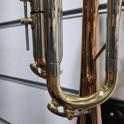 Close-up of a brass instrument, showing shiny gold and copper-toned metal valves, tubing, and bell against a light gray slatted wall background.
