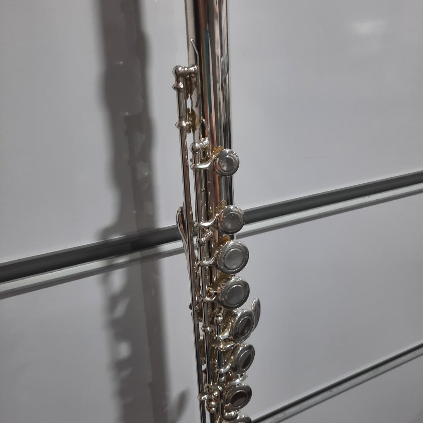 A close-up of a silver flute body with its key mechanism, photographed against a white paneled background.