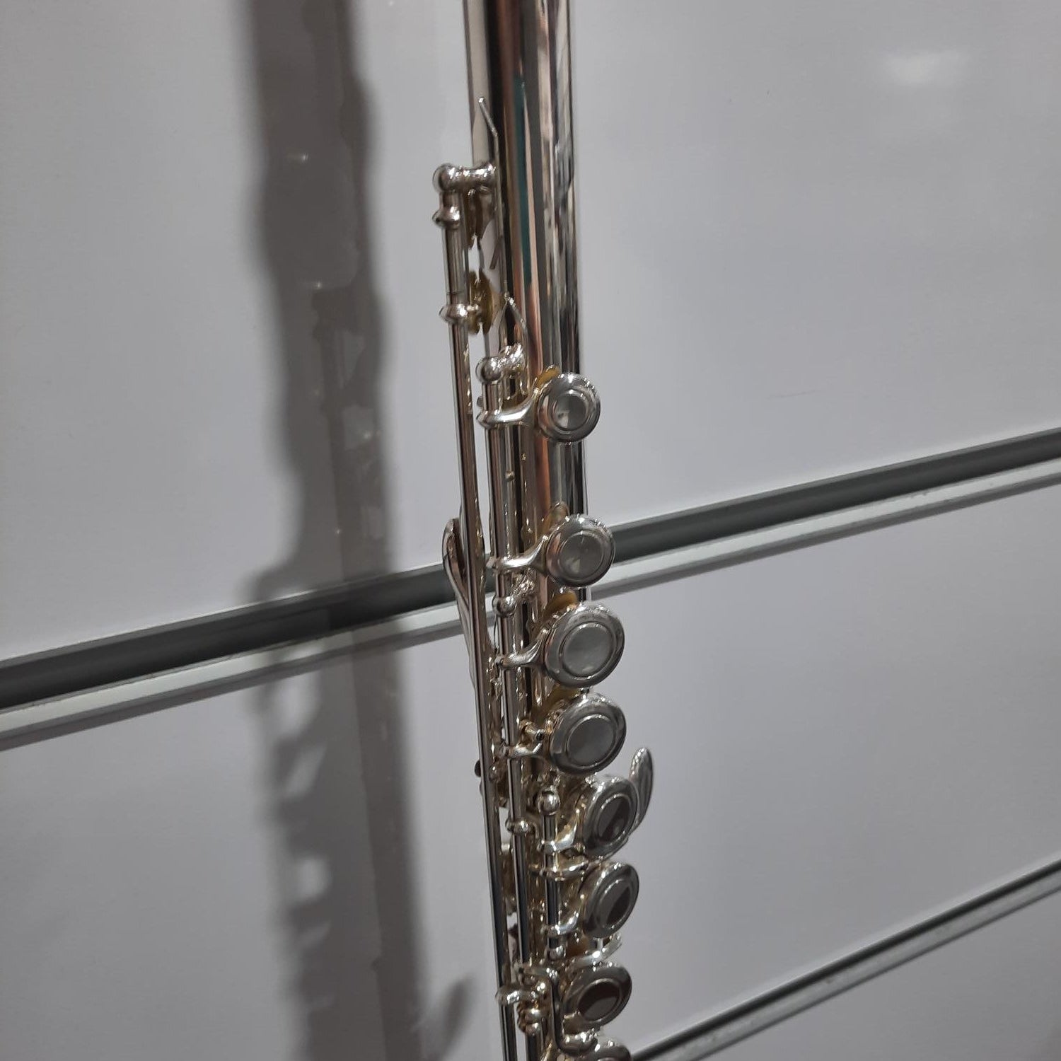 A close-up of a silver flute body with its key mechanism, photographed against a white paneled background.