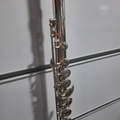 A close-up of a silver flute body with its key mechanism, photographed against a white paneled background.