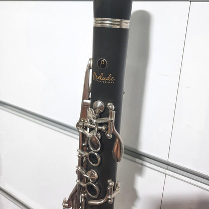 A black woodwind instrument, likely a clarinet, with silver metal keys and valves, standing vertically against a white tiled wall. The brand name 'Prelude' is visible on the body.